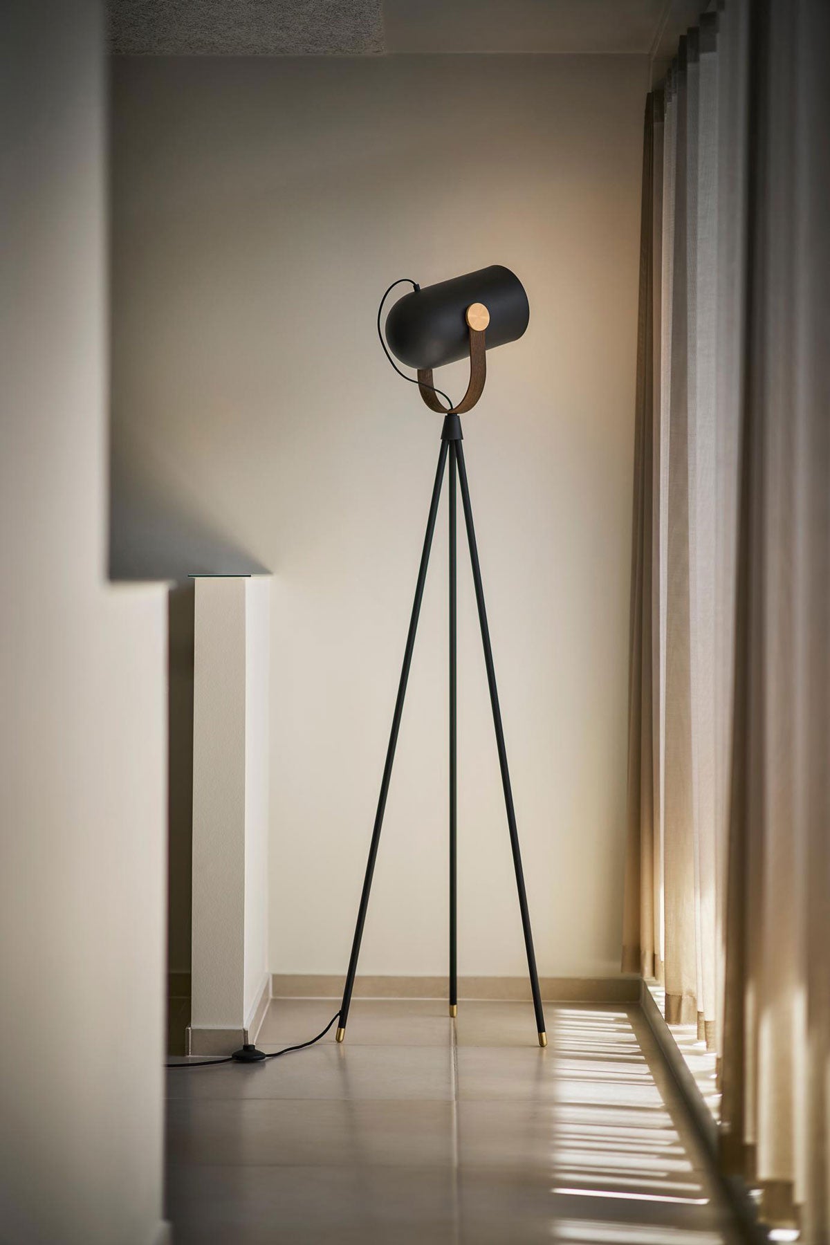 Carronade Golvlampa i rökt ek med mässingsdetalj framför fönster med soligt ljusinsläpp
