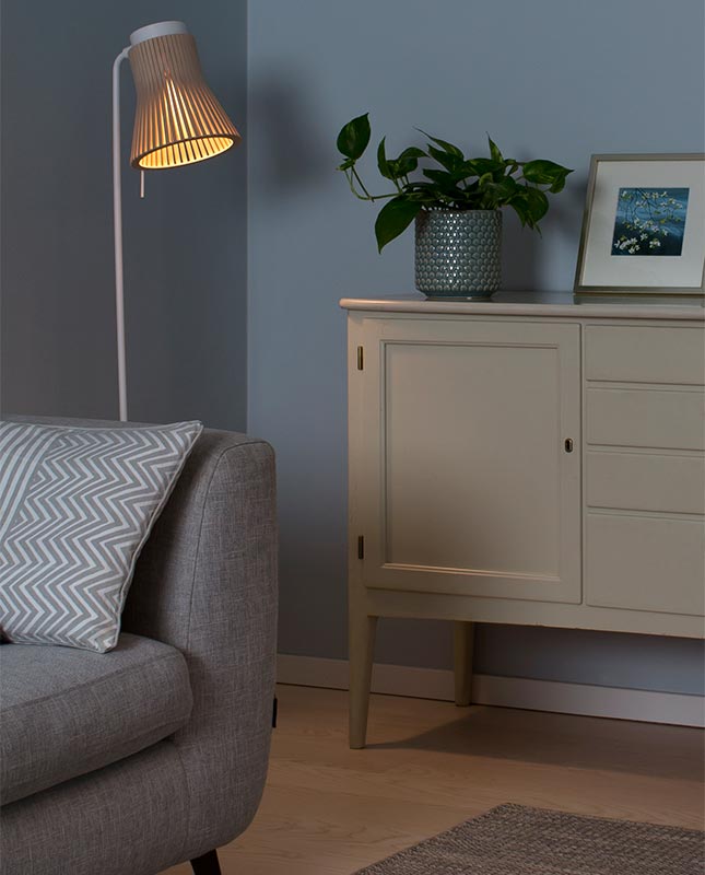 Petite 4610 Golvlampa Björk bredvid en skänk med en kruka och ett inramat fotografi